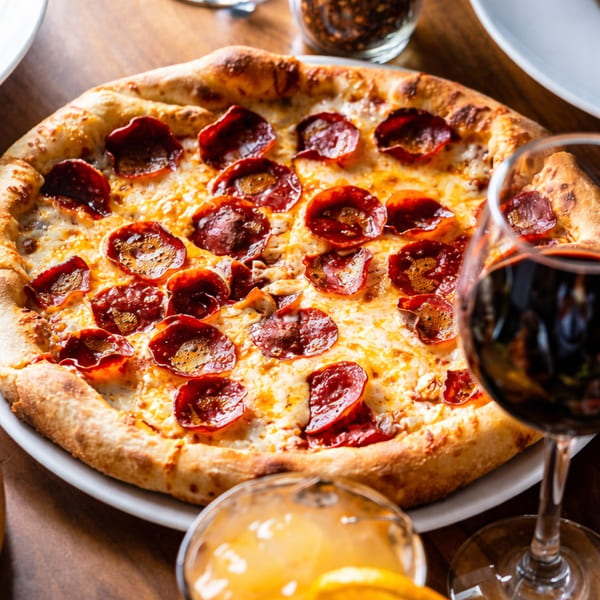 A pepperoni pizza with melted cheese and a golden crust, alongside glasses of red wine and orange juice.